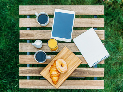 早上早餐绿色花园与法国牛角包,甜甜圈,咖啡杯,橙汁,平板电脑笔记簿木桌上背景图片