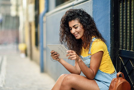 轻的阿拉伯女人户外看她的数字平板电脑非洲女孩穿着休闲服装城市背景卷发图片