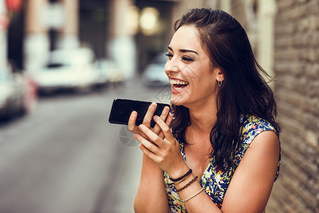 微笑的轻女人户外用她的智能手机录制语音音符城市背景下穿花裙的女孩技术图片