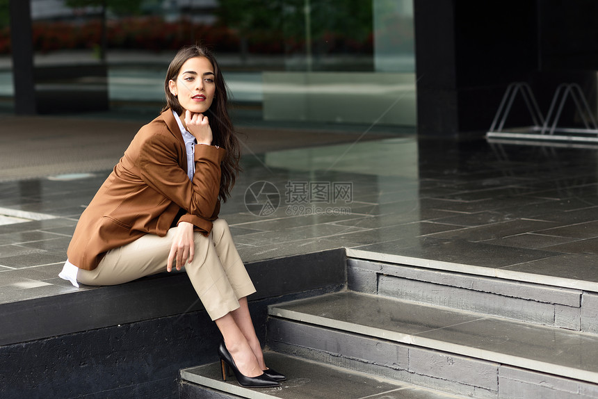 坐办公楼外的轻女商人穿正式衣服的漂亮女人轻女孩,棕色夹克裤子城市背景图片
