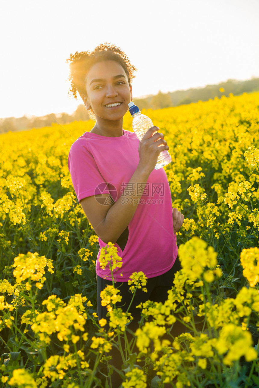 户外肖像美丽的快乐混合种族非裔美国女孩十几岁的女轻女子运动员瓶子里喝水,日落的黄色花朵金色的傍晚阳光图片