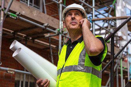 建筑工地上的男建筑工头工人建筑师持建筑计划,并手机上交谈背景图片