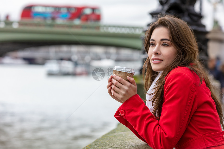 穿着红色外套的女孩轻女子威斯敏斯特桥旁边的次杯子里喝咖啡,背景伦敦英国大列颠的红色双层巴士图片