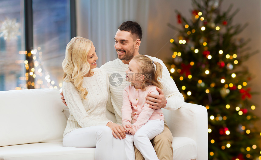 家庭,假期人的快乐的母亲,父亲小女儿坐沙发上,家里的诞树灯光背景幸福的家庭家里的诞树灯图片