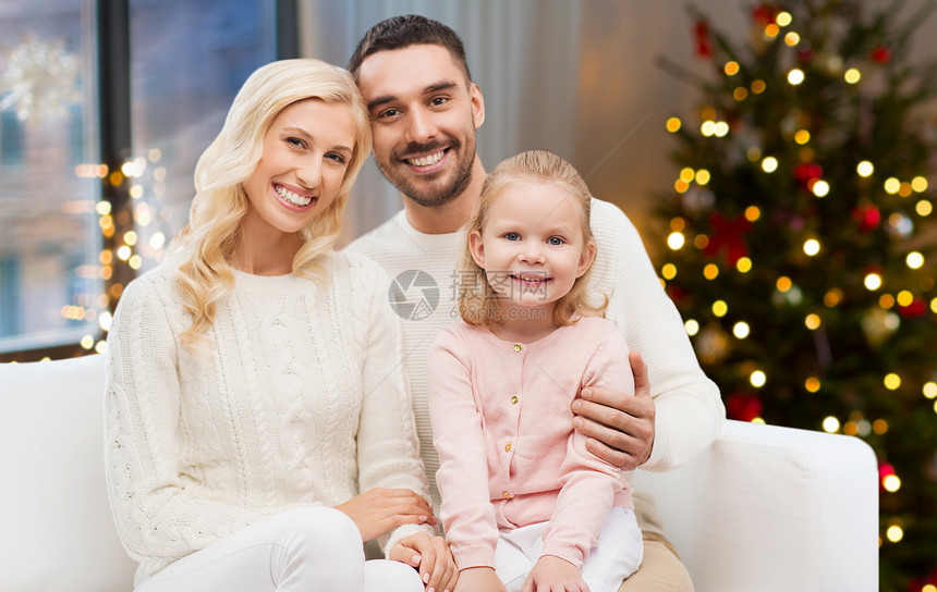 家庭,假期人的快乐的母亲,父亲小女儿坐沙发上,家里的诞树灯光背景幸福的家庭家里的诞树灯图片