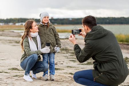 家庭休闲人的快乐的母亲父亲小女儿秋天的海滩上用智能手机拍照秋天的海滩上用智能手机拍摄家庭照片背景图片