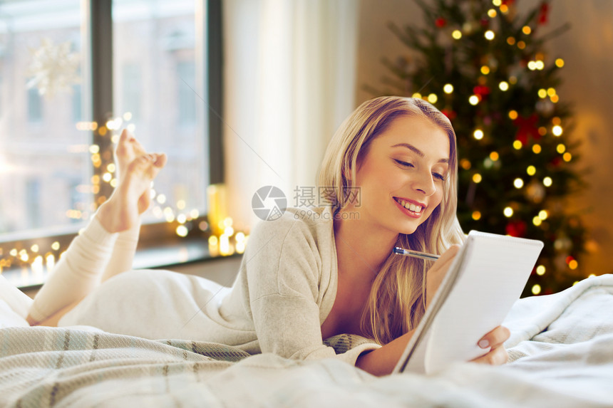 假期人们的快乐的轻女人写信笔记本家里的诞树灯光下床上列出愿望清单诞节家列愿望清单的女人图片