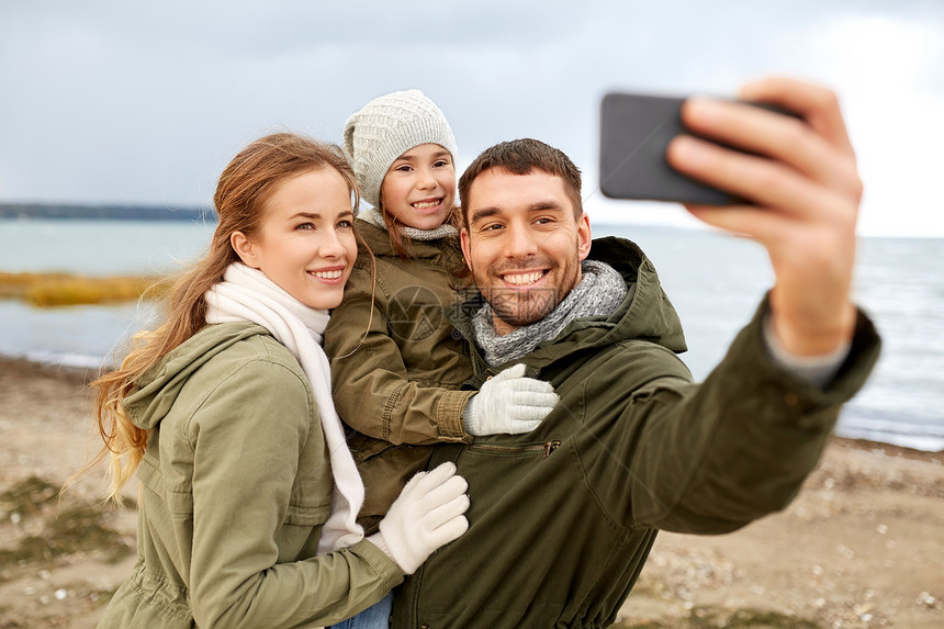 家庭休闲人的快乐的母亲父亲小女儿秋天的海滩上用智能手机自拍家人秋天的海滩上用智能手机自拍图片