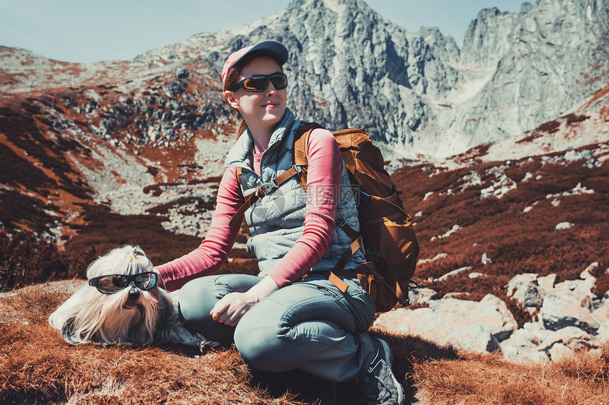 轻的女游客,带着狗太阳镜坐高山背景上秋天的白色红色图片