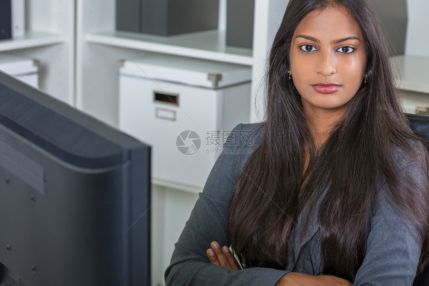 位美丽而权势的亚洲轻印度妇女办公室女商人的肖像,坐办公桌前,双臂交叉,用电脑折叠图片
