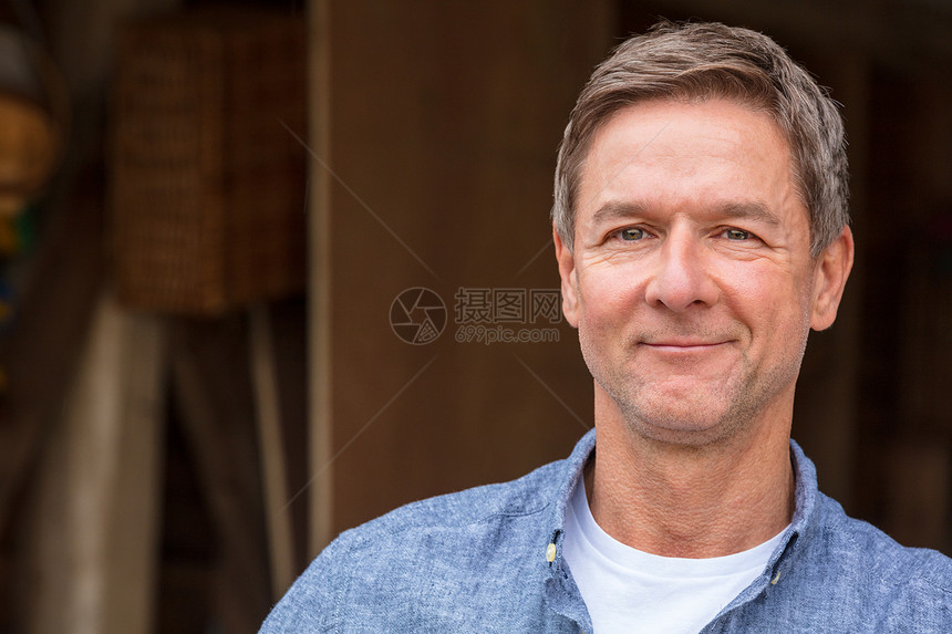 位迷人成功快乐的中男子的肖像照,他穿着件蓝色衬衫,靠车库谷仓的柱子上图片