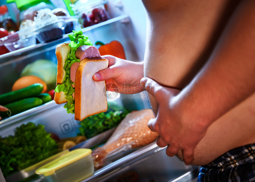 饥饿的胖子手里着个大三明治,站打开的冰箱旁边健康的食物图片