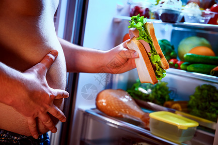 饥饿的胖子手里着个大三明治,站打开的冰箱旁边健康的食物背景图片