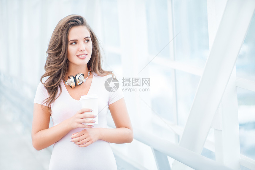 快乐的轻女人戴着音乐耳机,站桥上着咖啡杯,着城市背景向边看图片