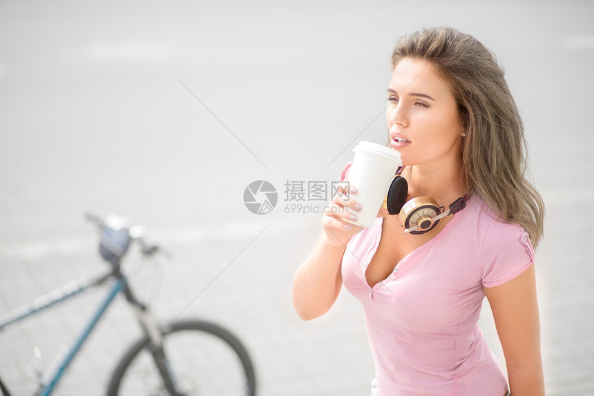 美丽的轻女人戴着音乐耳机脖子上,自行车道路背景下喝咖啡,个带走咖啡杯图片