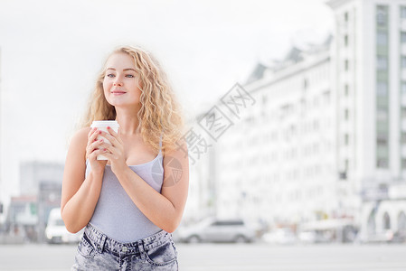 快乐的轻女人,长发卷曲,着个走的咖啡杯,微笑着城市交通背景下的相机前调情图片