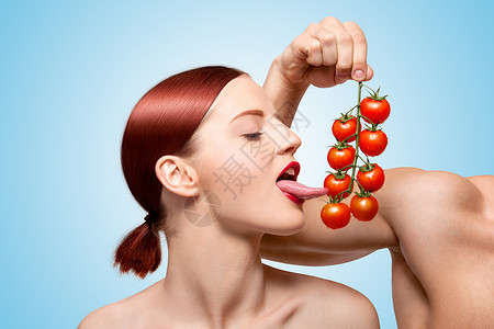 女舌头美丽的女孩用她诱人的舌头藤蔓上舔成熟的红色樱桃西红柿前戏与食物喂食蓝色背景背景