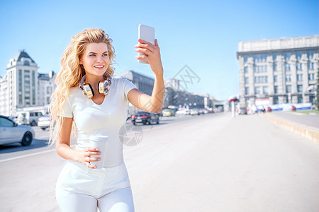 美丽的轻女人带着音乐耳机个带走咖啡杯,拍照自己,自拍城市背景图片