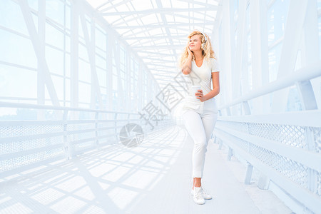 栏杆桥美丽的轻女人戴着音乐耳机,站桥上着咖啡杯,城市背景下摆姿势背景