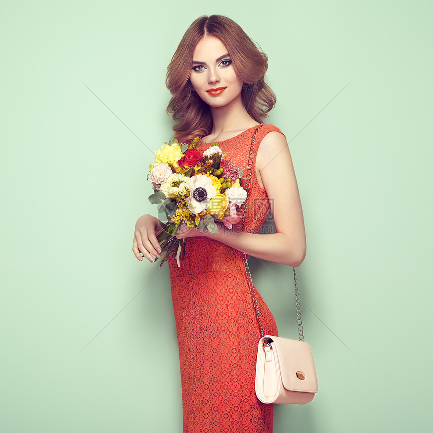 穿着优雅红色连衣裙的金发轻女人女孩用手提包摆绿色背景上珠宝发型带着春天花的女士时尚照片图片