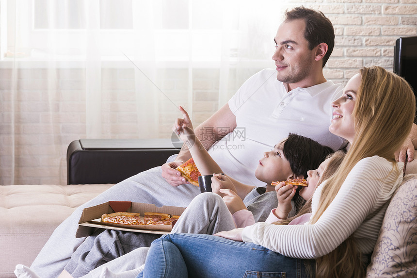 轻的父母他们的孩子家里坐沙发上看电视,吃披萨微笑图片