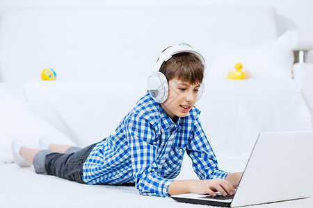 孩子享受闲暇时间学龄男孩躺地板上,耳机听笔记本电脑上的音乐图片