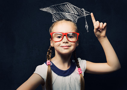 聪明的女学生戴红眼镜的天才女孩戴着硕士帽靠近黑板图片