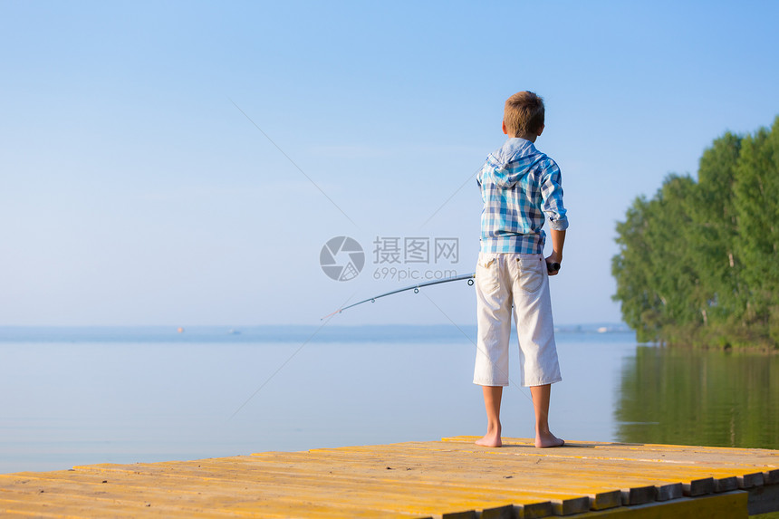 穿着蓝色衬衫的男孩站海边的码头上,着鱼竿穿着蓝色衬衫的男孩站馅饼上图片