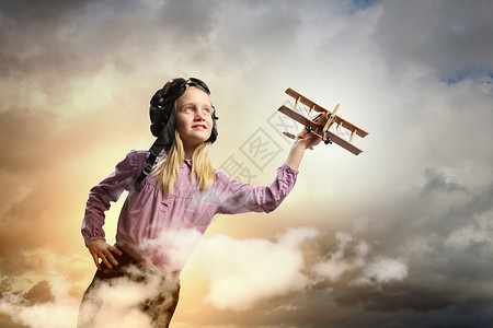 戴着飞行员帽子的小女孩飞行员头盔中的小女孩云背景下玩玩具飞机的形象图片