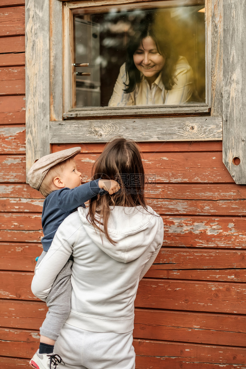 农村的场景,岁的小男孩戴着平帽,妈妈户外看着小屋的窗户,祖母图片