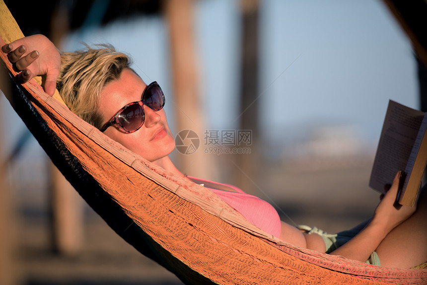 放松的女人躺沙滩上的吊床上,边看书边享受日落图片
