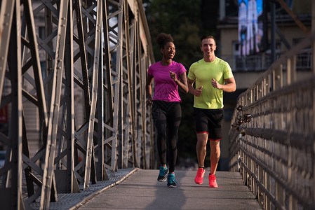 健康的轻多民族夫妇清晨城市的桥上慢跑,背景日出图片