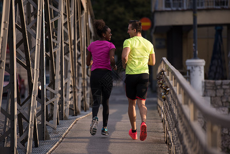 健康的轻多民族夫妇清晨城市的桥上慢跑,背景日出图片