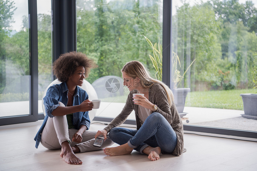 两个轻的笑着的多民族妇女坐靠近窗户的地板上,边喝咖啡,边豪华的家里用智能手机喝咖啡图片