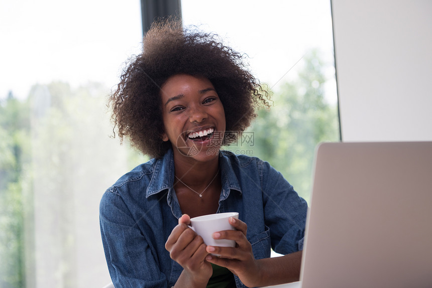 轻的非裔美国妇女微笑着坐明亮的窗户附近,看着桌子上打开的笔记本电脑,着白色的杯子她豪华的家图片