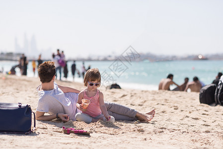 酷的轻妈妈她的小女孩玩着桶装满沙子的太阳镜,阳光明媚的天海滩上放松图片