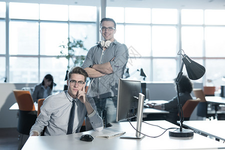 两个商人用计算机为下次会议准备,并与背景中的同事讨论想法图片