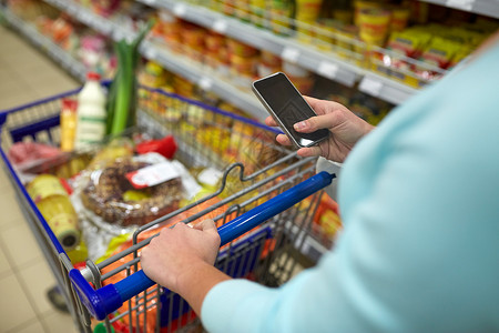 消费主义,技术人的妇女与智能手机,购物车手推车购买食品杂货店超市图片