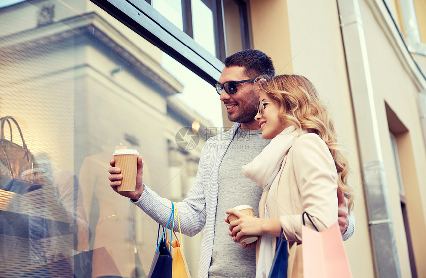 销售,消费主义人们的幸福的夫妇与购物袋咖啡纸杯看着城市的商店窗口城市里购物袋咖啡的幸福夫妇图片