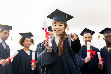 选择董事会教育毕业人的群快乐的国际学生穿着灰浆板学士服,文凭指向你快乐的学生用文凭指着你背景