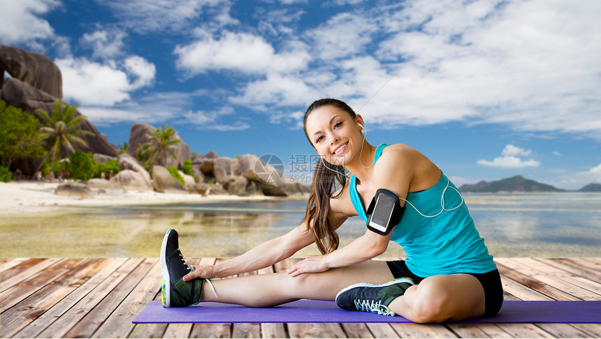 健身,运动,锻炼,技术人的微笑的女人戴着与智能手机耳机伸展腿垫子上的异国情调热带海滩背景智能手机的女人垫图片