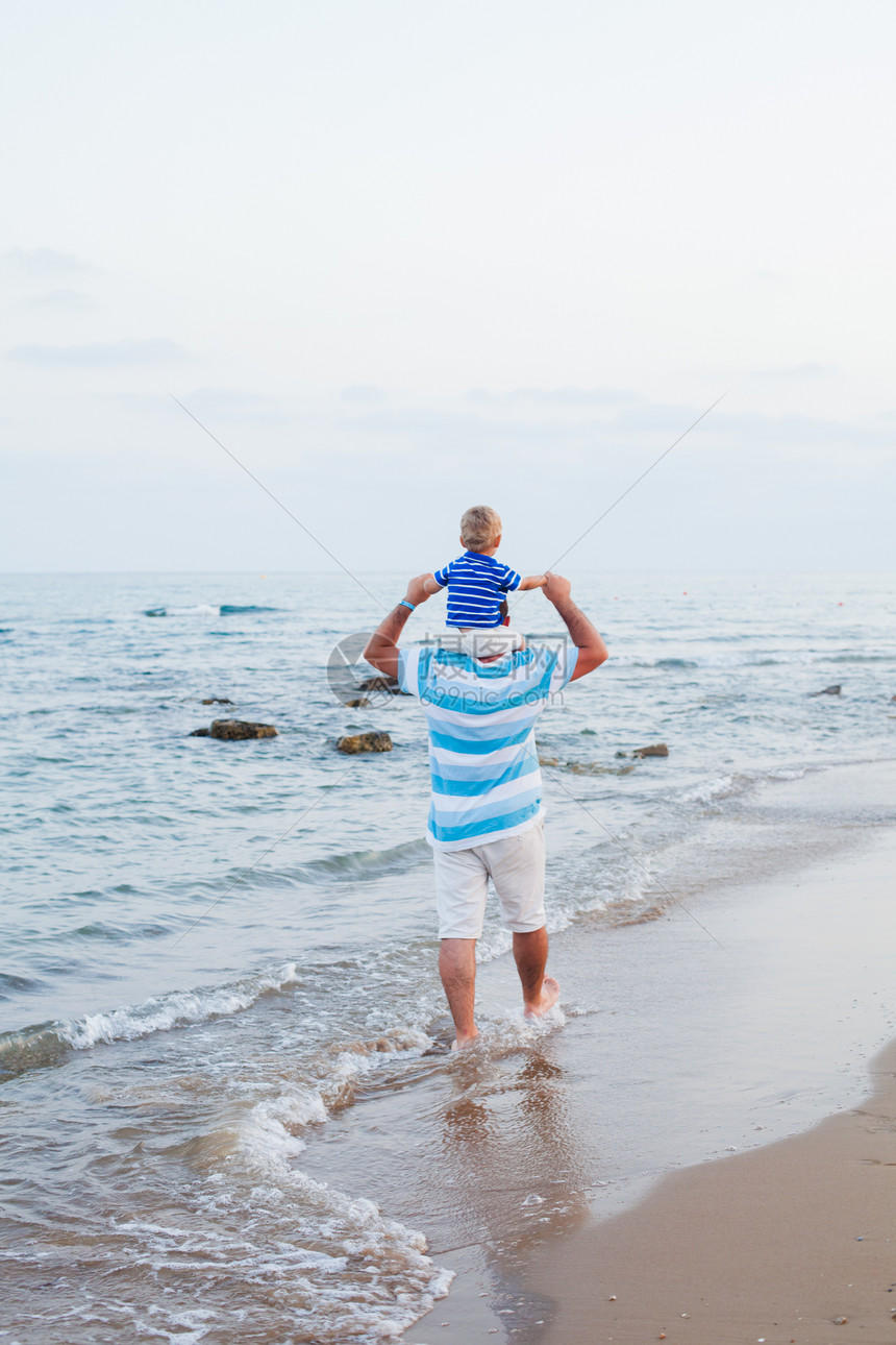 轻的父亲他的小儿子夏天花时间户外沿着沙滩散步儿子坐父亲的肩膀上玩得很开心图片