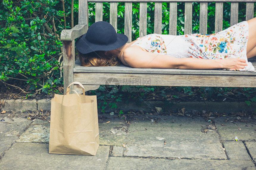个轻的女人穿着件夏装顶帽子,阳光明媚的日子里公园的长凳上放松,她旁边个纸袋图片