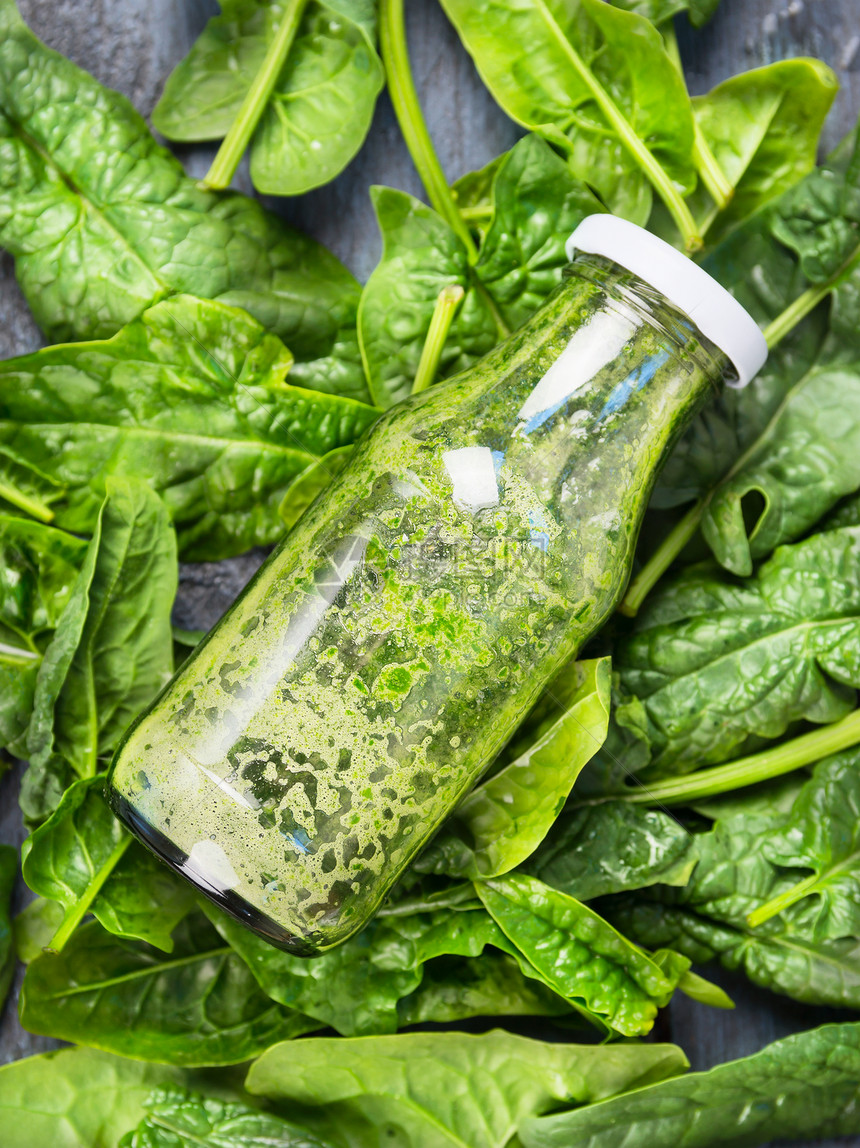 瓶中的绿色冰沙位于新鲜菠菜背景上,顶部视图图片
