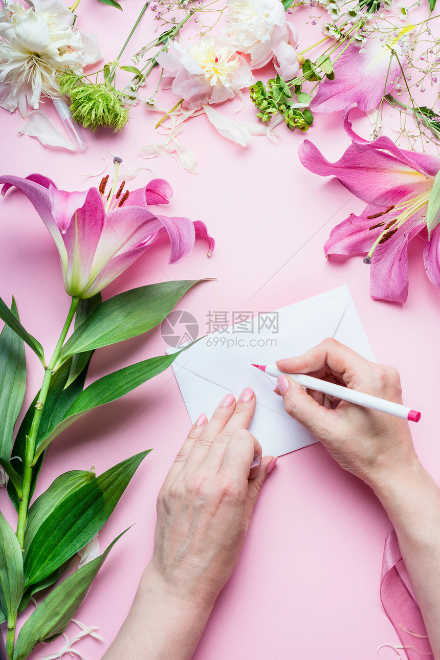 女的手用铅笔写贺卡粉红色桌子背景上的空白信封上,百合花花店装饰设备,顶部视图创意邀请假期图片