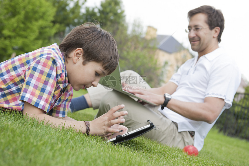 父子俩用笔记本电脑平板电脑躺草地上图片