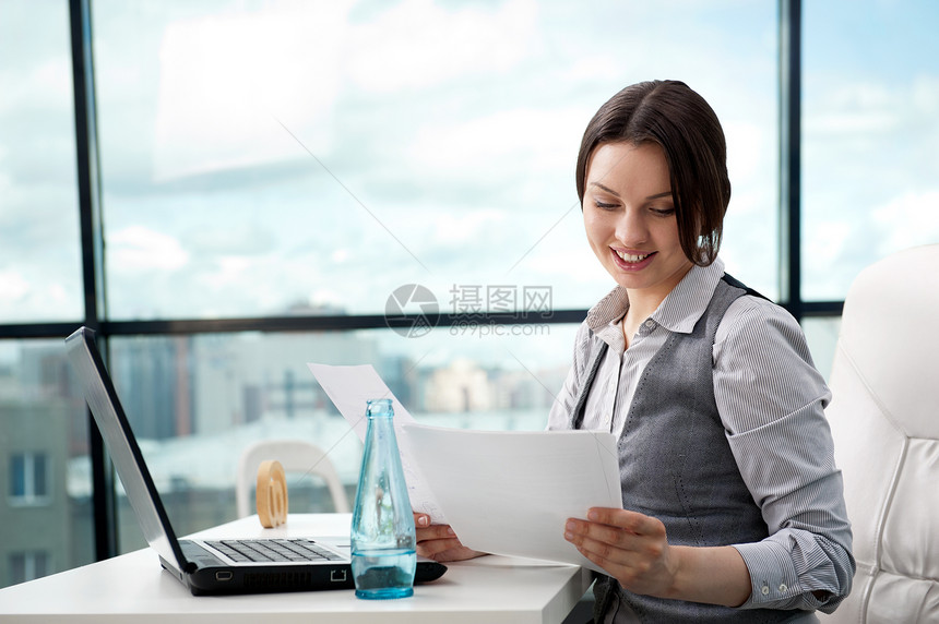 美丽的商业妇女看着她抱怀里的文件,同时她的办公室工作的电脑图片