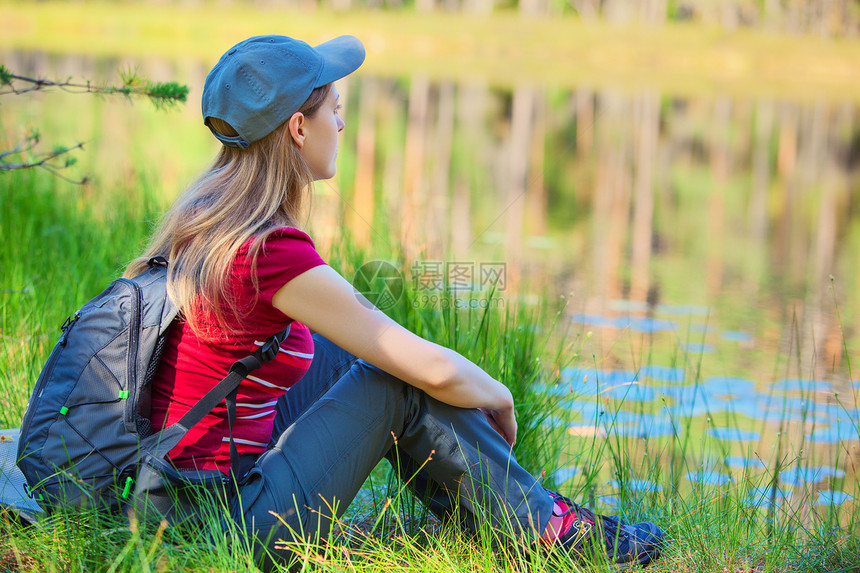 轻的女游客坐池塘海岸的草地上图片