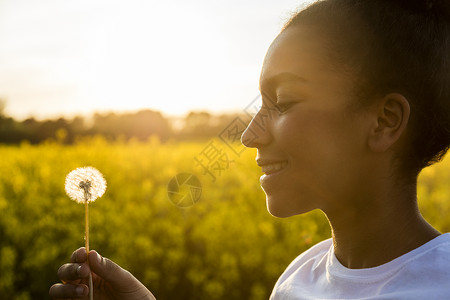 户外肖像美丽的快乐混合种族非裔美国女孩十几岁的女轻女子微笑金色的夕阳灯光蒲公英时钟花背景