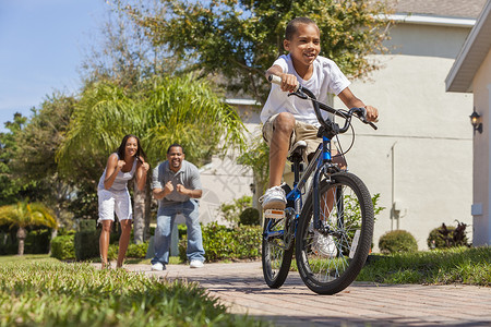 个轻的非裔美国人家庭,男孩骑着自行车,他快乐而兴奋的父母他身后给予鼓励图片